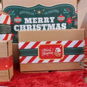 Kotak hadiah bertema Natal dengan label “Merry Christmas” berwarna merah dan hijau, dihiasi ilustrasi Santa dan rusa di atas permukaan karpet merah, menciptakan suasana hangat dan festive.