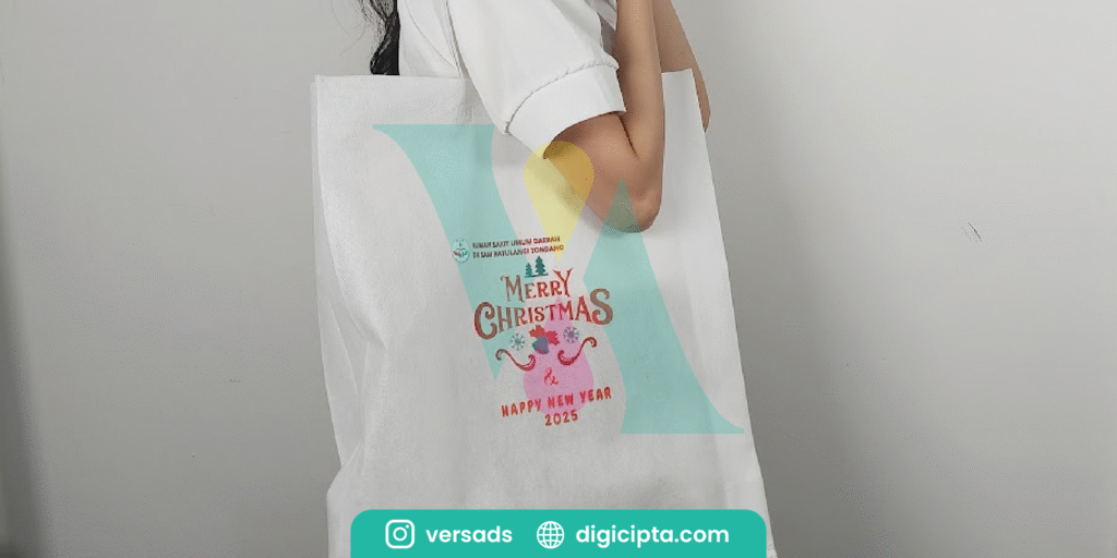 Perempuan mengenakan totebag putih bertuliskan “Merry Christmas & Happy New Year 2025” dengan desain grafis bertema Natal berwarna cerah dan minimalis.