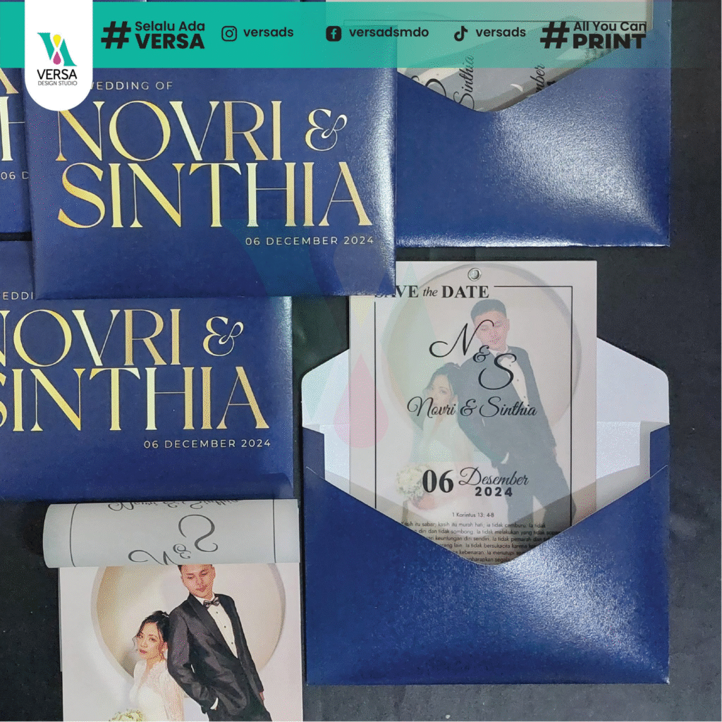 kumpulan undangan pernikahan elegan berwarna biru tua bertuliskan “Novri & Sinthia 06 December 2024” dengan font emas mengkilap, lengkap dengan amplop matching dan kartu save the date bergambar pasangan pengantin berpakaian formal, hasil cetak dari Versa Design Studio Manado.